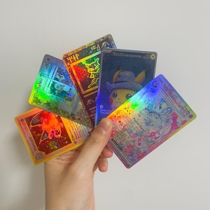 Puede incluir: Una mano sosteniendo una colecci&oacute;n de cartas coleccionables hologr&aacute;ficas de Pok&eacute;mon. Las cartas presentan varios personajes, incluyendo Pikachu, Charizard y Sylveon, con colores vibrantes y detalles de l&aacute;mina. Las cartas son rectangulares y tienen texto y n&uacute;meros.