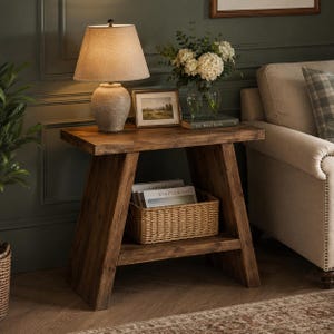 May include: A wooden side table with an A-frame base and a lower shelf containing a woven wicker basket. The tabletop holds a textured ceramic lamp with a beige fabric shade, a small framed landscape print, and a glass vase with white flowers.