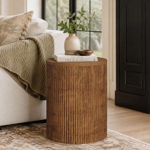 May include: A cylindrical side table made of medium-brown wood with a vertical fluted texture. On top of the table sit two stacked white books with the text "THIS IS HOME" and "LIVE BEAUTIFULLY" printed on the spines, a small wooden bowl, and a vase.