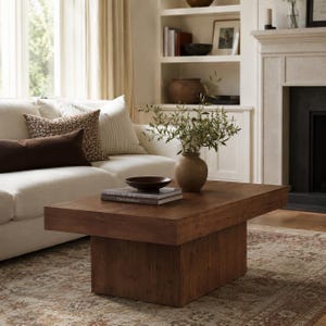 May include: A rectangular wooden coffee table with a thick top and a solid block base. The table is finished in a medium brown wood grain and holds a small dark bowl, two stacked books, and a round ceramic vase with green leafy branches.