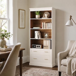 May include: A tall white bookcase featuring four open shelves with a dark wood-toned back panel and two bottom drawers with gold-toned horizontal handles. The unit has a decorative fluted top molding and is filled with various books and decor.