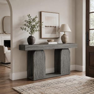 May include: A gray wooden console table with a thick rectangular top and two arched support bases. On the table sits a dark ceramic vase with green foliage, a stack of books, a small bowl, and a table lamp with a beige fabric shade.