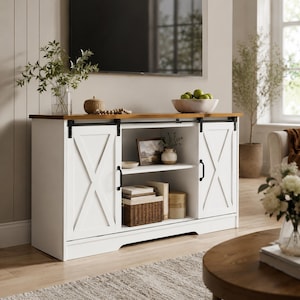 May include: A white and brown TV stand with sliding barn doors. The stand has a wooden top and a white base with an X-shaped design on the doors. The stand has a shelf inside and is decorated with a bowl of green apples.