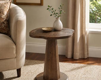 May include: A round wooden side table with a natural wood grain finish on the top surface, featuring a pedestal base and a circular bottom support. A small white ceramic vase with green leaves and a small gold bowl sit on the table top.