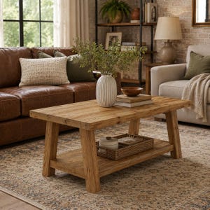 May include: A rectangular wooden coffee table with a lower shelf, featuring a light brown wood grain finish. On the tabletop sits a white ribbed ceramic vase with green foliage, a small wooden bowl, and two stacked books. A woven tray sits below.