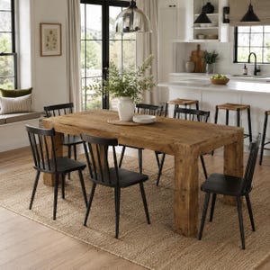 May include: A rectangular wooden dining table with a natural finish and thick legs, surrounded by six black spindle-back chairs. A white vase containing green foliage sits in the center of the table, which is placed on a large woven area rug.