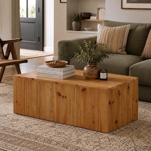 May include: A rectangular coffee table crafted from light brown wood with a visible grain and knot pattern. The table features a solid, box-like construction with a flat top surface, providing a sturdy base for decorative items and books.