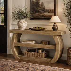 May include: A light brown wooden console table with a curved base and two shelves. The top surface holds a white textured vase with greenery, a wooden bowl with green spheres, and a lamp with a pleated shade. Books and a basket are on the shelves.