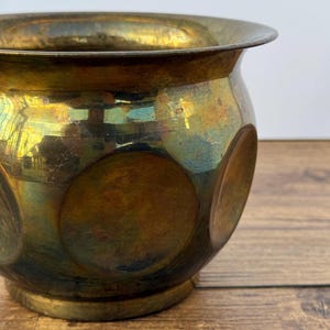 Puede incluir: Un cuenco redondo de latón con un borde ancho y acampanado y un acabado pulido. La superficie exterior presenta una serie de grandes hendiduras circulares dispuestas alrededor del cuerpo, mostrando oxidación natural y tonos metálicos dorados.