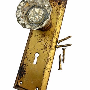 May include: A vintage door knob set featuring a clear, faceted glass knob attached to a rectangular, weathered brass backplate with a central keyhole. The set includes four small brass screws positioned to the side of the backplate.