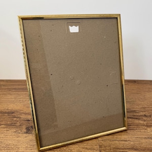 May include: A rectangular picture frame with a thin metallic gold border. The frame features a brown cardboard backing with a centred, notched metal hanger at the top for wall mounting. The design is simple, functional, and clean-lined.