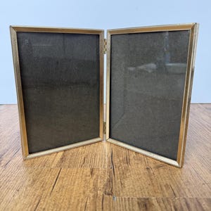 Puede incluir: Un marco de fotos doble con bisagras que presenta dos paneles rectangulares con bordes finos de metal dorado. El marco tiene un detalle de borde texturizado con cuentas a lo largo del perímetro interior de cada lado y está unido por dos pequeñas bisagras.