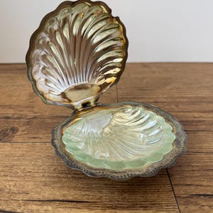 May include: A hinged clam shell shaped trinket box with a metallic gold finish on the exterior and interior lid. The base features a translucent pale green glass insert moulded into a ribbed shell pattern with a scalloped edge and metal rim.