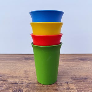 Op de afbeelding: Een stapel van vier plastic bekers in een verticale opstelling. De bovenste beker is blauw, de tweede is geel, de derde is rood en de onderste beker is groen. Elke beker heeft een gladde, matte afwerking met een licht uitlopende rand.