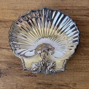 May include: A silver-toned metal dish shaped like a scallop shell with a polished, ribbed interior surface. The base features a decorative relief ornament depicting a cluster of grapes and vine leaves with a textured, matte finish.