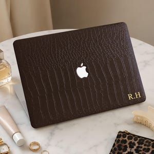 May include: A dark brown laptop cover with a textured crocodile skin pattern and the gold initials R.H. printed in the bottom right corner. The cover features a central cutout displaying the illuminated white Apple logo on the laptop lid.