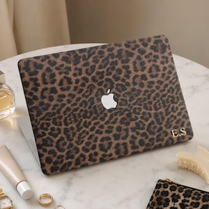May include: A laptop skin featuring a brown and black leopard print pattern with a white Apple logo cutout in the center and the gold initials E.S printed in the bottom right corner, displayed on a closed laptop computer.
