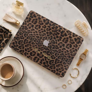 Può includere: Una skin per laptop con motivo leopardato marrone e nero e il nome Alexis Phillip in caratteri dorati. La skin è applicata su un laptop con il logo Apple argentato visibile, circondato da vari accessori personali.