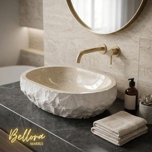 May include: An oval-shaped beige stone vessel sink with a rough, natural rock-hewn exterior and a smooth interior basin. The sink is paired with a wall-mounted brushed gold faucet and handle, set against a light-coloured stone wall and dark countertop.