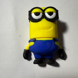 May include: A yellow Minion figurine with black goggles, black gloves, and black shoes. The Minion wears blue overalls. The figurine is 3D printed and stands on a white surface.