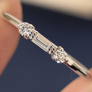 Op de afbeelding: Een dunne zilverkleurige ring met een centrale baguette-geslepen steen geflankeerd door twee ronde briljant-geslepen stenen. De metalen band is glad en gepolijst en toont een strak ontwerp met drie heldere, gefacetteerde edelstenen op een rij.