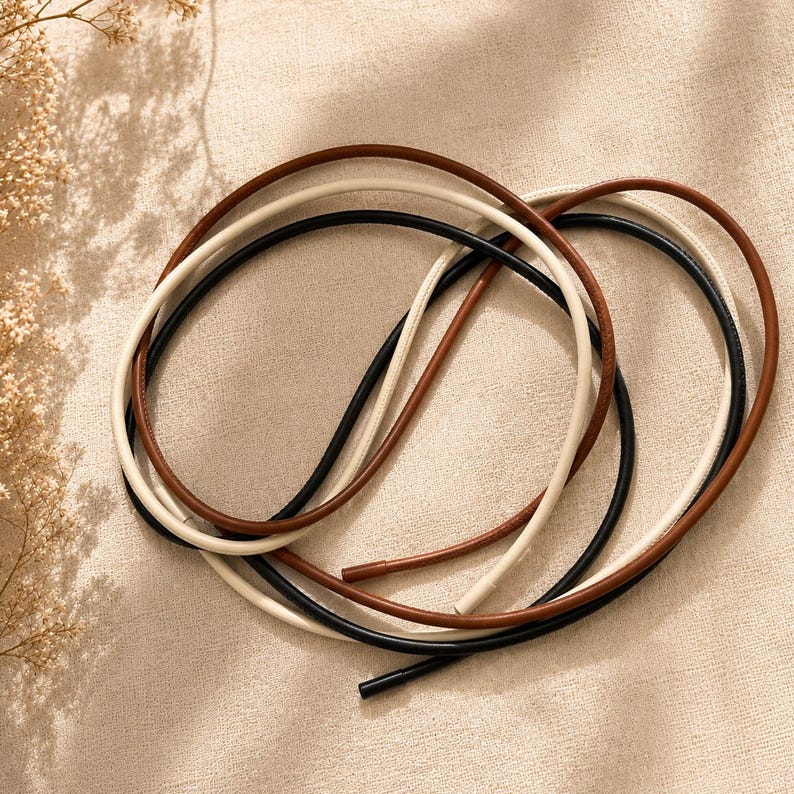 Op de afbeelding: Drie lange, dunne, flexibele koorden in zwart, cr&egrave;me en bruin. Elk koord heeft een glad, afgerond oppervlak en een kleine, cilindrische metalen dop aan het uiteinde. De koorden zijn in een los, overlappend cirkelvormig patroon gerangschikt.