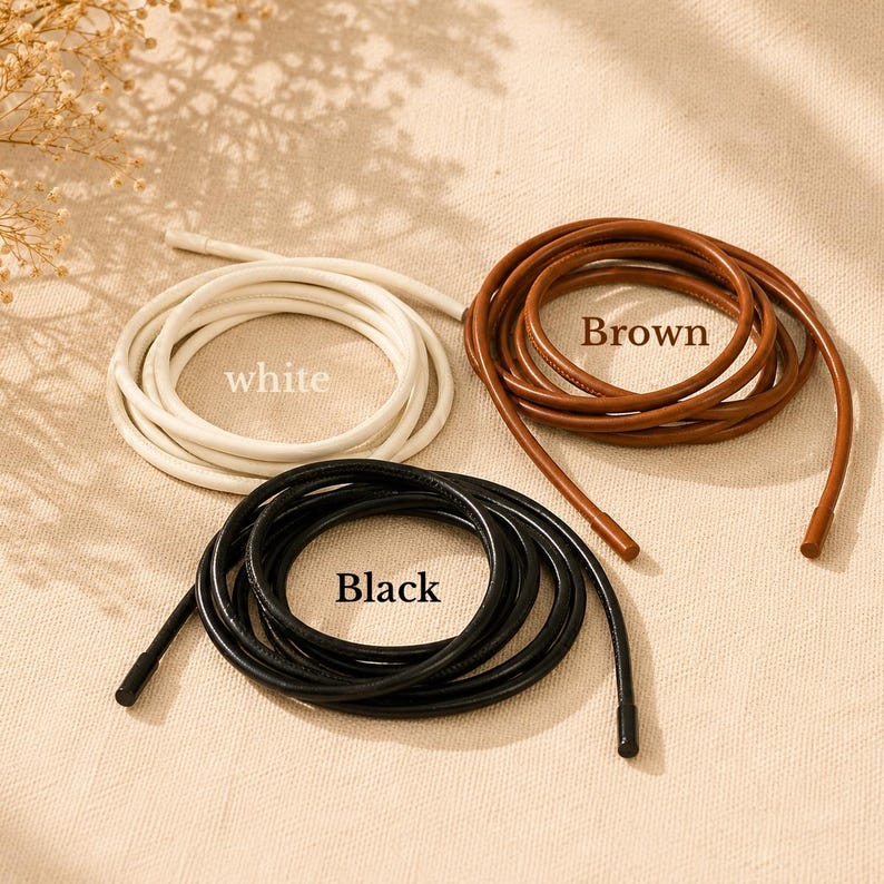 Op de afbeelding: Drie opgerolde koorden in wit, bruin en zwart, elk met gladde, afgeronde oppervlakken en bijpassende eindkappen. Het witte koord is gelabeld met de tekst white, het bruine koord met Brown en het zwarte koord met de tekst Black.