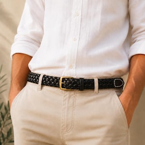 Può includere: Una cintura intrecciata in pelle nera con fibbia in metallo color ottone, indossata nei passanti di un pantalone beige. La cintura presenta una trama intrecciata e un'estremità a punta con un dettaglio di cucitura bianca vicino alla punta.