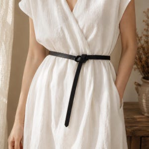 May include: A black leather belt cinches the waist of a white dress. The belt is tied in a knot, with a long, slender end hanging down. The dress has a V-neck and a simple, elegant design. The overall aesthetic is minimalist and chic.
