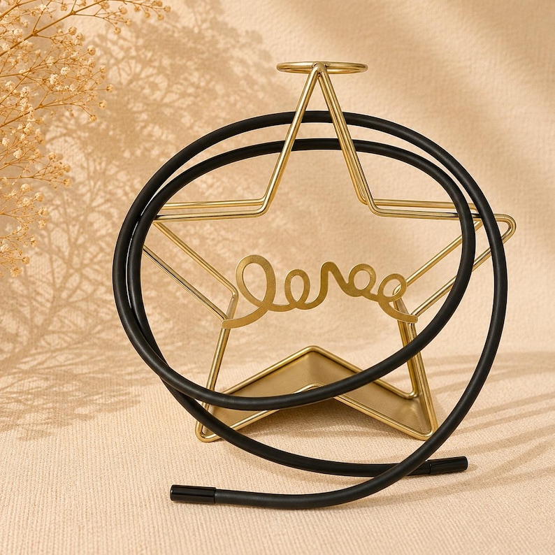 Op de afbeelding: Een goudkleurige metalen ster-vormige displaystandaard met het woord love in cursief schrift in het midden verwerkt. Een glad zwart koord is over het frame gedrapeerd, dat een kleine cirkelvormige lus aan de bovenkant en een stevige voet heeft.
