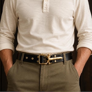 Peut inclure: Une ceinture en cuir noir dotée d'une boucle en laiton poli avec un mécanisme de fermeture à crochet. La ceinture est fixée par des rivets en laiton et est portée sur un pantalon vert olive avec un t-shirt henley à manches longues crème.