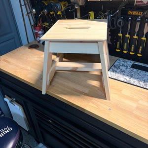 May include: A light-colored wooden step stool featuring a rectangular top with a centered handle cutout, four angled legs, and horizontal support braces connecting the legs. The stool has a smooth, unfinished natural wood grain texture.