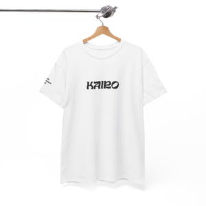 Peut inclure: T-shirt blanc avec le mot "KAIRO" imprimé en noir sur la poitrine. Le t-shirt est suspendu à un cintre en bois. Le texte est imprimé verticalement sur la manche gauche. Le t-shirt est en matière souple.