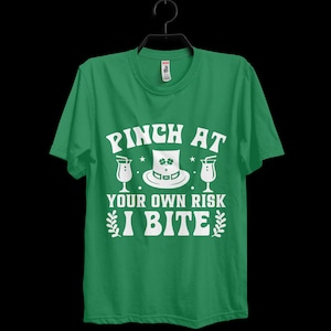 May include: A bright green short-sleeve t-shirt featuring white graphic text that reads PINCH AT YOUR OWN RISK I BITE. The design includes a central leprechaun hat with a shamrock, flanked by two cocktail glasses and small decorative leaf sprigs.