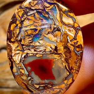 Könnte beinhalten: Ovale Opal-Edelstein mit einer lebendigen Farbpalette. Der Stein zeigt eine Mischung aus Gold-, Braun-, Blau- und Rottönen mit komplizierten Mustern und Texturen. Die polierte Oberfläche reflektiert das Licht und zeigt die einzigartige Struktur des Opals.