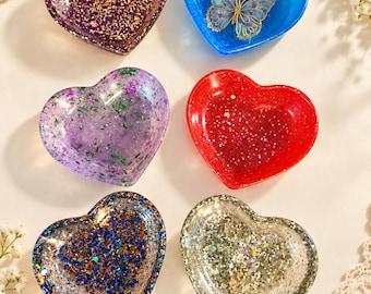 Puede incluir: Seis cuencos de resina en forma de corazón con varios acabados de purpurina. Un cuenco es morado y dorado, uno azul con una mariposa incrustada, uno morado claro con escamas verdes, uno rojo, uno azul y plateado, y uno plateado.