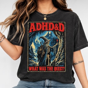 Può includere: Una t-shirt grigio antracite con una stampa grafica di uno scheletro mago in una grotta. Il testo in alto recita ADHD&D in rosso, e il testo in basso recita WHAT WAS THE QUEST? in rosso. L'illustrazione include toni blu e gialli.