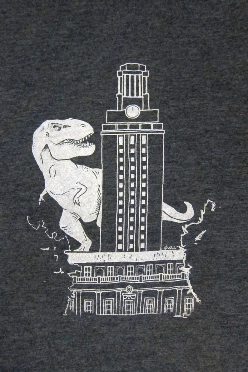 May include: A white graphic print on a dark grey t-shirt shows a Tyrannosaurus Rex dinosaur standing on top of the University of Texas at Austin's iconic tower, the clock tower.