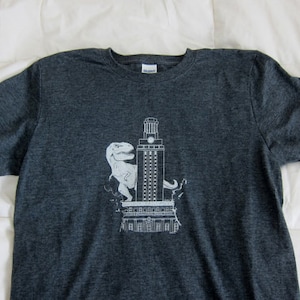 May include: A dark grey t-shirt featuring a white graphic of a dinosaur standing in front of a building. The building has a clock tower and the text "University of Texas at Austin" is printed on the shirt.