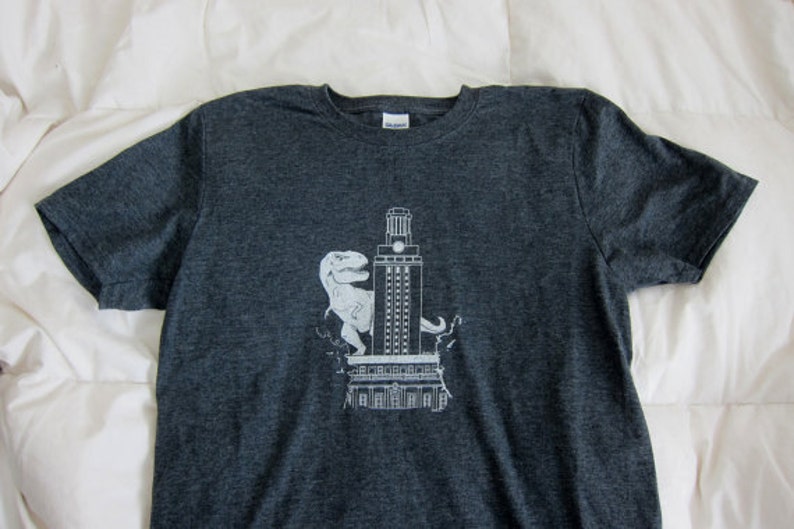 May include: A dark grey t-shirt featuring a white graphic of a dinosaur standing in front of a building. The building has a clock tower and the text "University of Texas at Austin" is printed on the shirt.