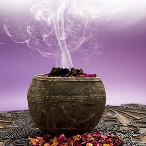 May include: A dark brown, round bowl with a carved design sits on a patterned surface. The bowl is filled with dried rose petals and incense, and smoke rises from the incense.