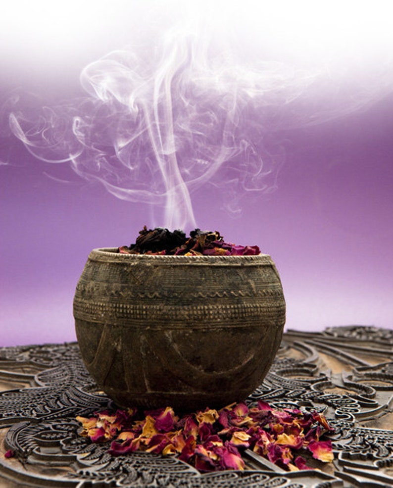 May include: A dark brown, round bowl with a carved design sits on a patterned surface. The bowl is filled with dried rose petals and incense, and smoke rises from the incense.
