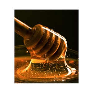 May include: A wooden honey dipper with honey dripping from it into a glass bowl. The honey is a golden amber colour and is thick and viscous.