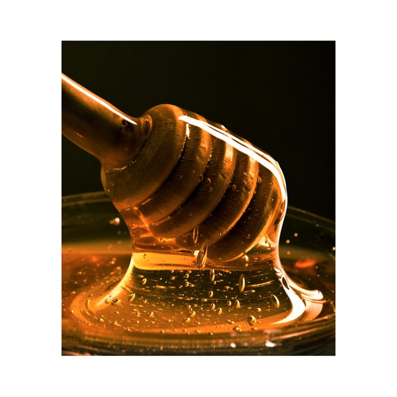 May include: A wooden honey dipper with honey dripping from it into a glass bowl. The honey is a golden amber colour and is thick and viscous.