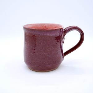 May include: A handmade ceramic mug in a deep burgundy color with a matching handle. The interior of the mug is a lighter shade of red. The mug has a simple, classic design, perfect for enjoying coffee or tea.
