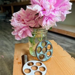 Op de afbeelding: Twee ronde keramische bloemenhouders met elk zes gaten, één met een glanzend blauwgroen glazuur en de andere met een gladde witte afwerking, geplaatst bij een doorzichtige glazen pot gevuld met water en twee grote, levendige roze pioenrozen.