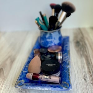 Puede incluir: Una bandeja de cerámica azul contiene brochas de maquillaje, esponjas y varios recipientes cosméticos. Una pequeña taza de cerámica azul y blanca contiene los pinceles. La bandeja es rectangular con una superficie texturizada y detalles dorados.