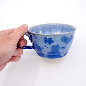 May include: A blue ceramic teacup with a handle, decorated with darker blue floral patterns. The cup has a wide rim and is held by a hand against a white background. The cup is approximately 4 inches in diameter.