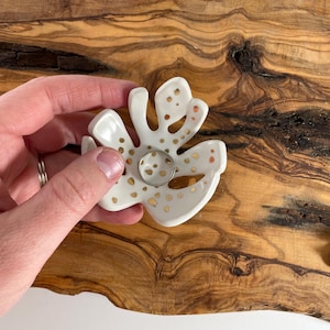 May include: White ceramic ring holder in a floral design with gold accents. The ring holder is held by a hand and is placed on a wooden surface. The ring holder has a circular center for holding rings.