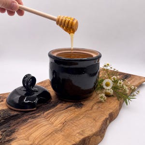 Puede incluir: Un tarro de miel de cerámica negra con tapa, lleno de miel. Un cucharón de miel de madera se sostiene sobre el tarro, con miel goteando. El tarro está sobre una tabla de madera para servir con flores.